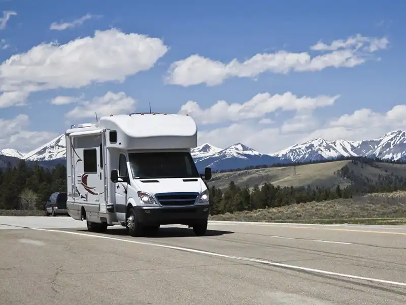 White motorhome driving on a highway with snow-capped mountains and blue sky in the background, trees on the side.