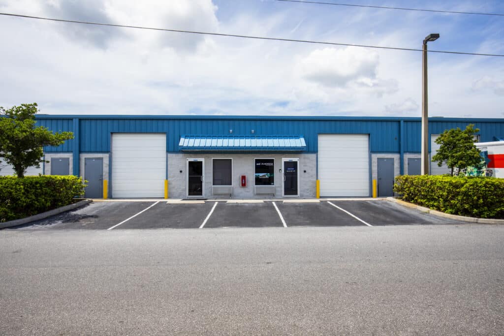 IFB 25-272-066 Blue industrial warehouse with white loading doors and a central entrance, small blue awning, and an empty parking lot in front.