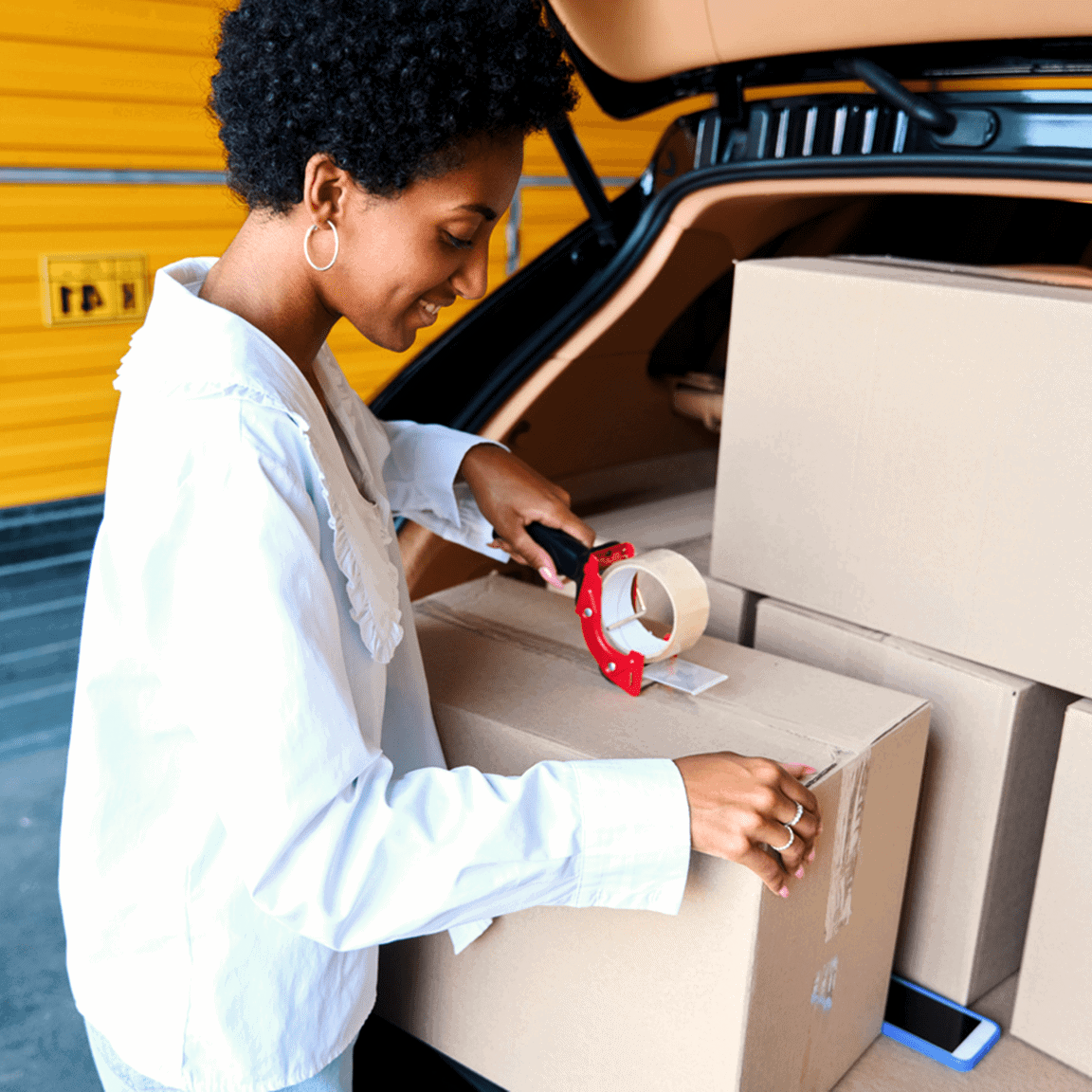 Young woman taping up a box in the back of her car, standing outside of a storage unit.