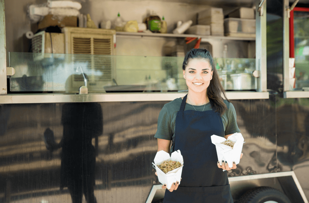Woman in an apron stands in front of her food truck holding takeout containers