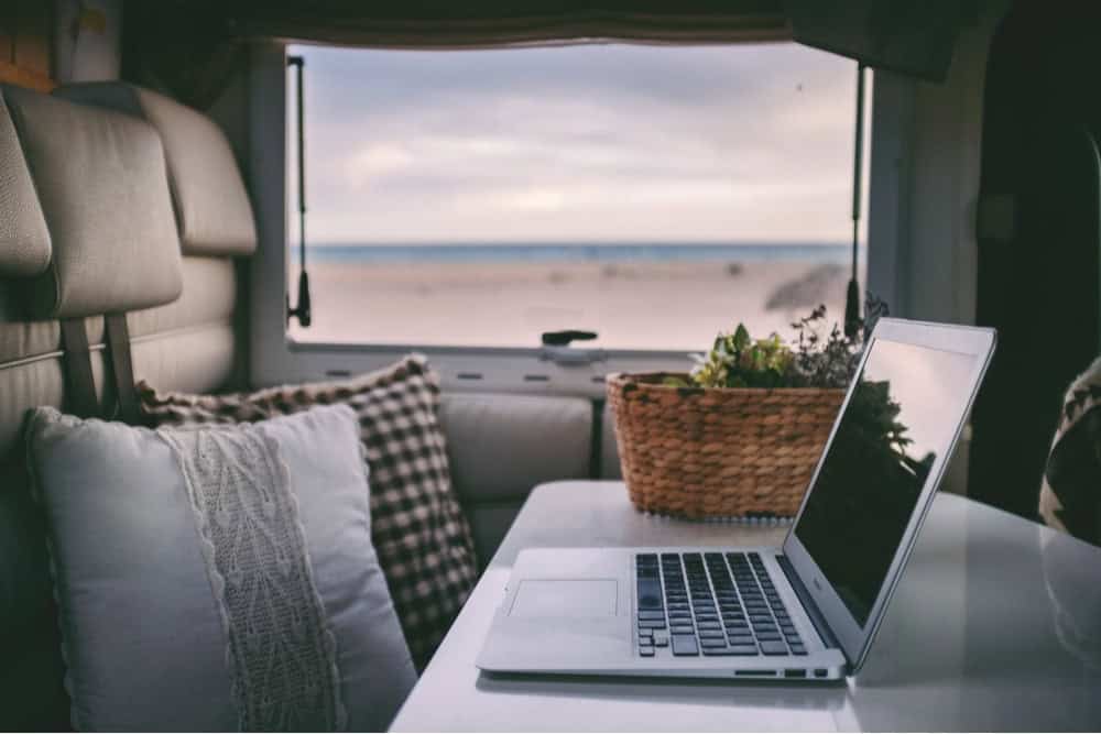 Laptop set up on RV dinette. RV window overlooks a calm beach.