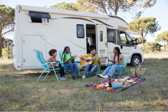 Family of four gathered around a picnic blanket outside of their RV; Father playing guitar.
