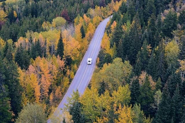 RV seen from far above, driving down a road in the fall, bordered by dense forest