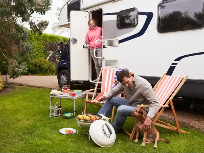 Man grilling on a grill outside a parked RV while a woman stands in the RV doorway, and a dog sits near striped deck chairs.