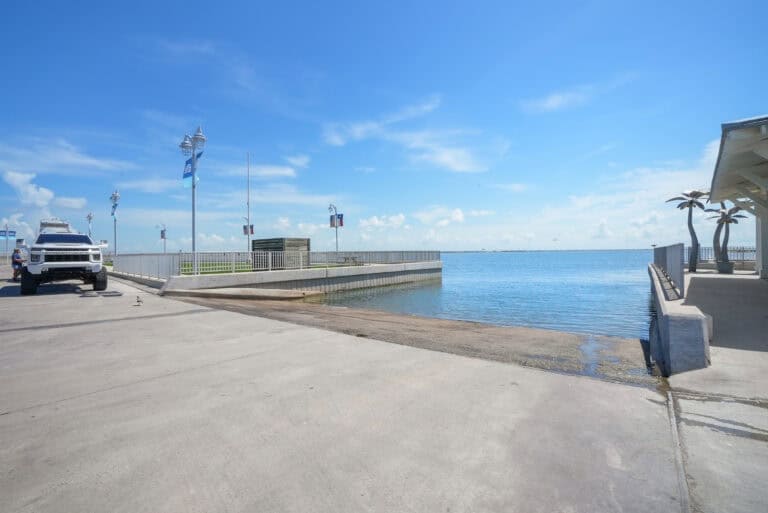 Bright sunny seaside with clear blue sky and calm water at a coastal pier, featuring palm trees and flagpoles.