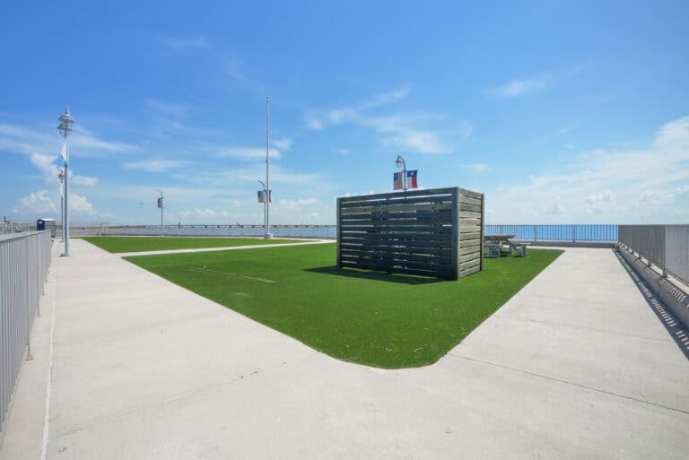 Bright outdoor terrace with artificial grass, wooden privacy wall, and ocean views under a blue sky.