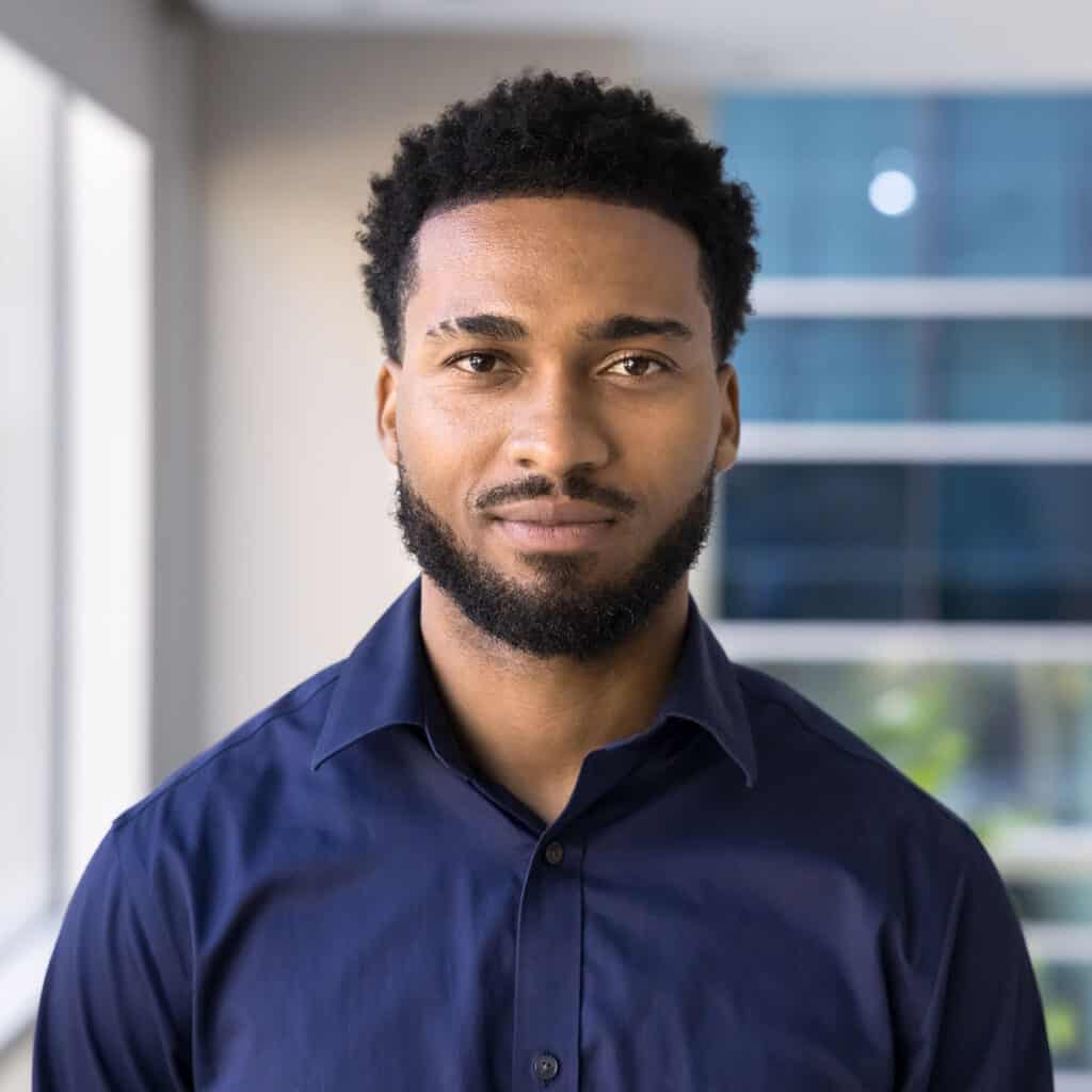 join_the_outrig_team Portrait of a man with short curly hair and a beard, wearing a navy button-down shirt, standing indoors by a window.