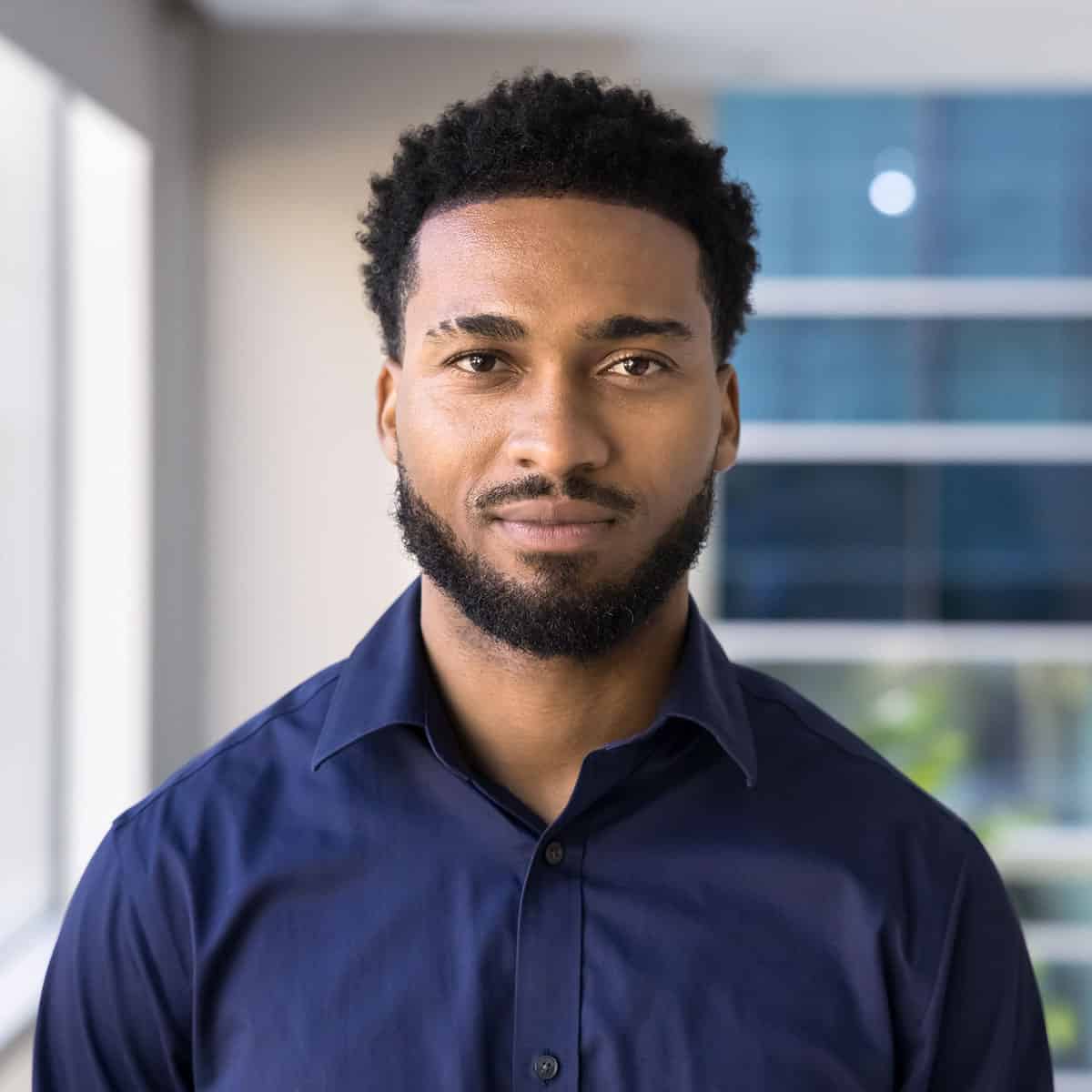 Portrait of a man with short curly hair and a beard, wearing a navy button-down shirt, standing indoors by a window.